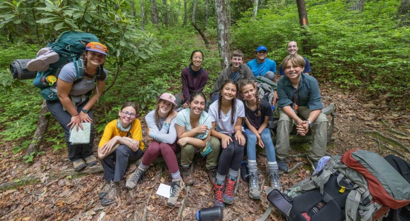 A group of young people sit together in a wooded area and smile for a group photo.